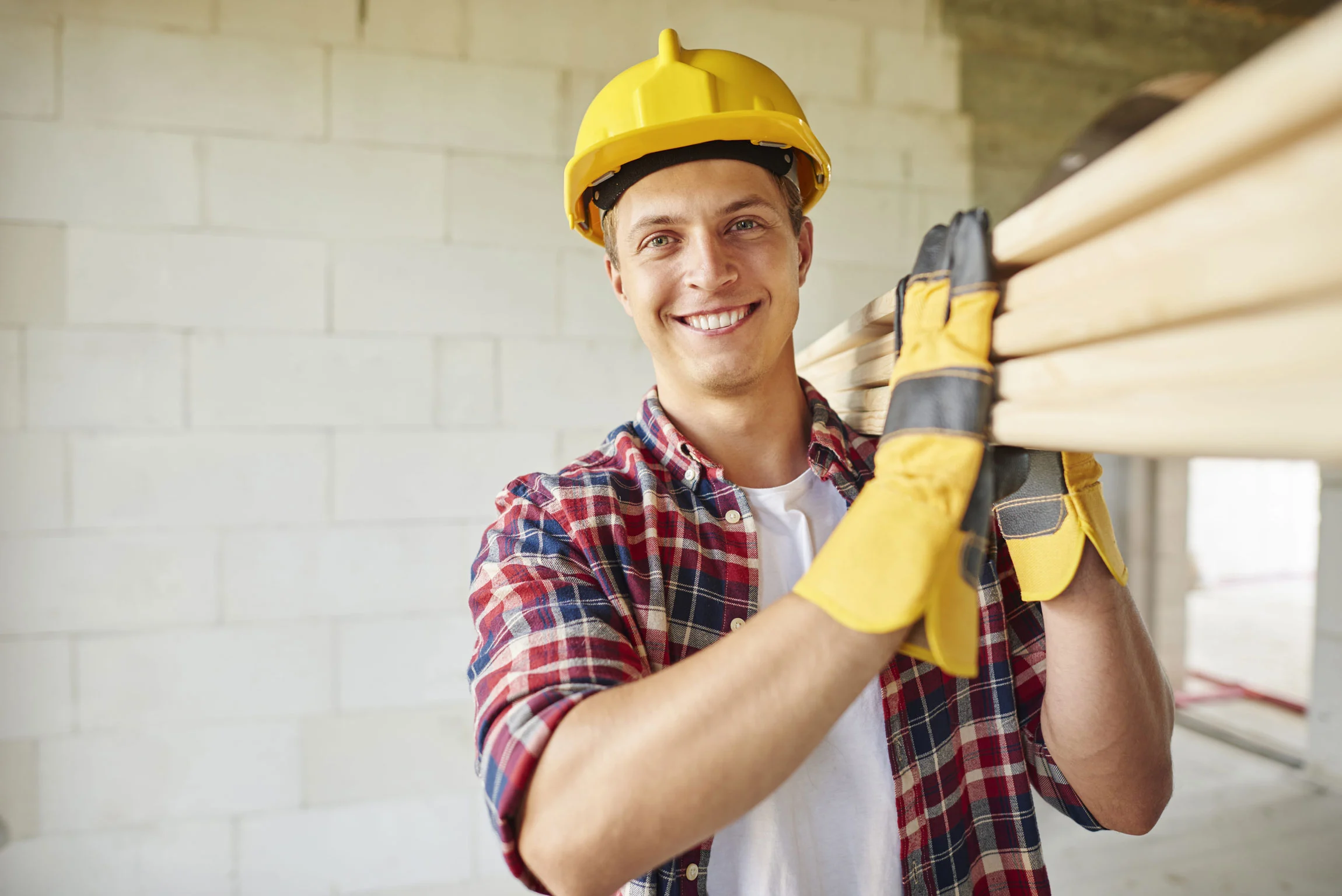 Handwerker mit Schutzhelm trägt Holzbohlen auf der Schulter durch einen Rohbau